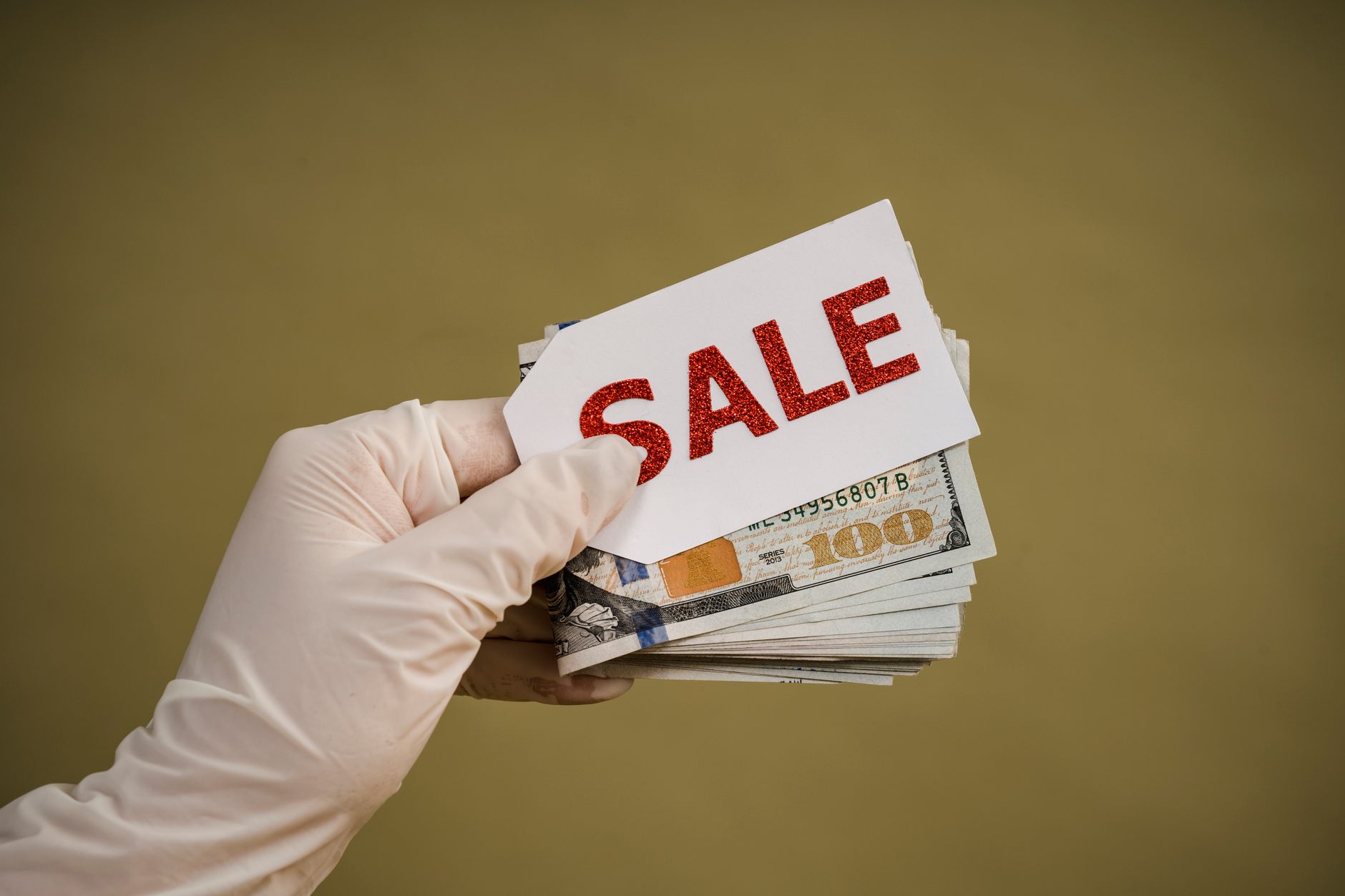 A hand wearing latex gloves holds a stack of dollar bills with a sale sign, symbolizing discount or cash transaction.