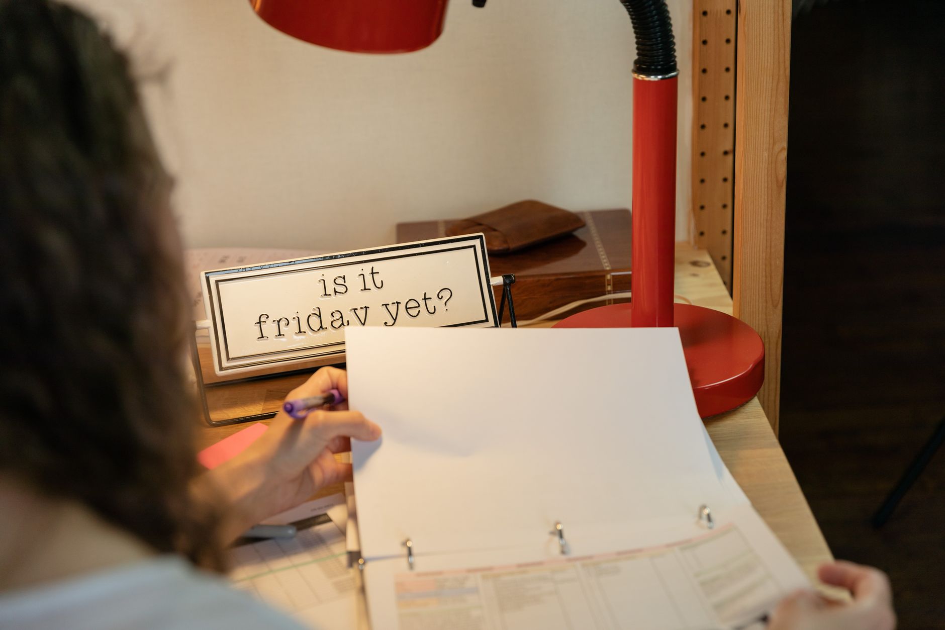 Cozy home office setup with red desk lamp, sign 'Is it Friday yet?', and person looking at documents.