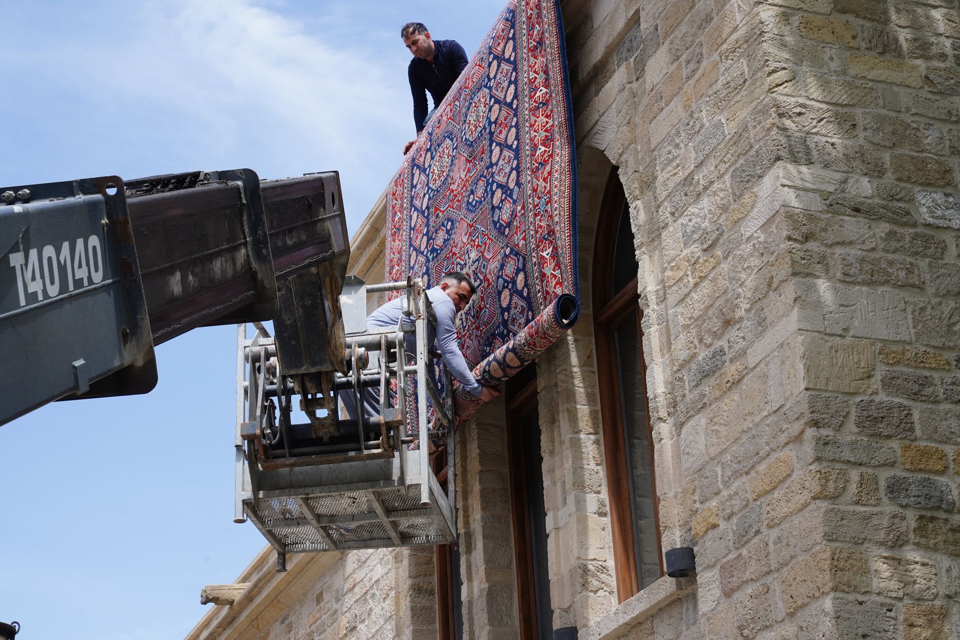 Workers use a lift to install a large patterned carpet on a stone building facade.